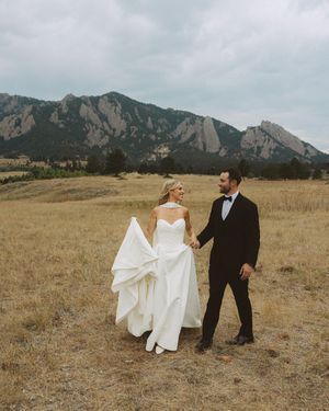Bride and groom holding hands in golden field with Colorado Flatirons mountain backdrop on their wedding day.