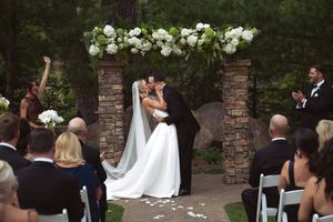 Bride and groom share first kiss at outdoor wedding ceremony under floral arch with white hydrangeas, guests applauding.
