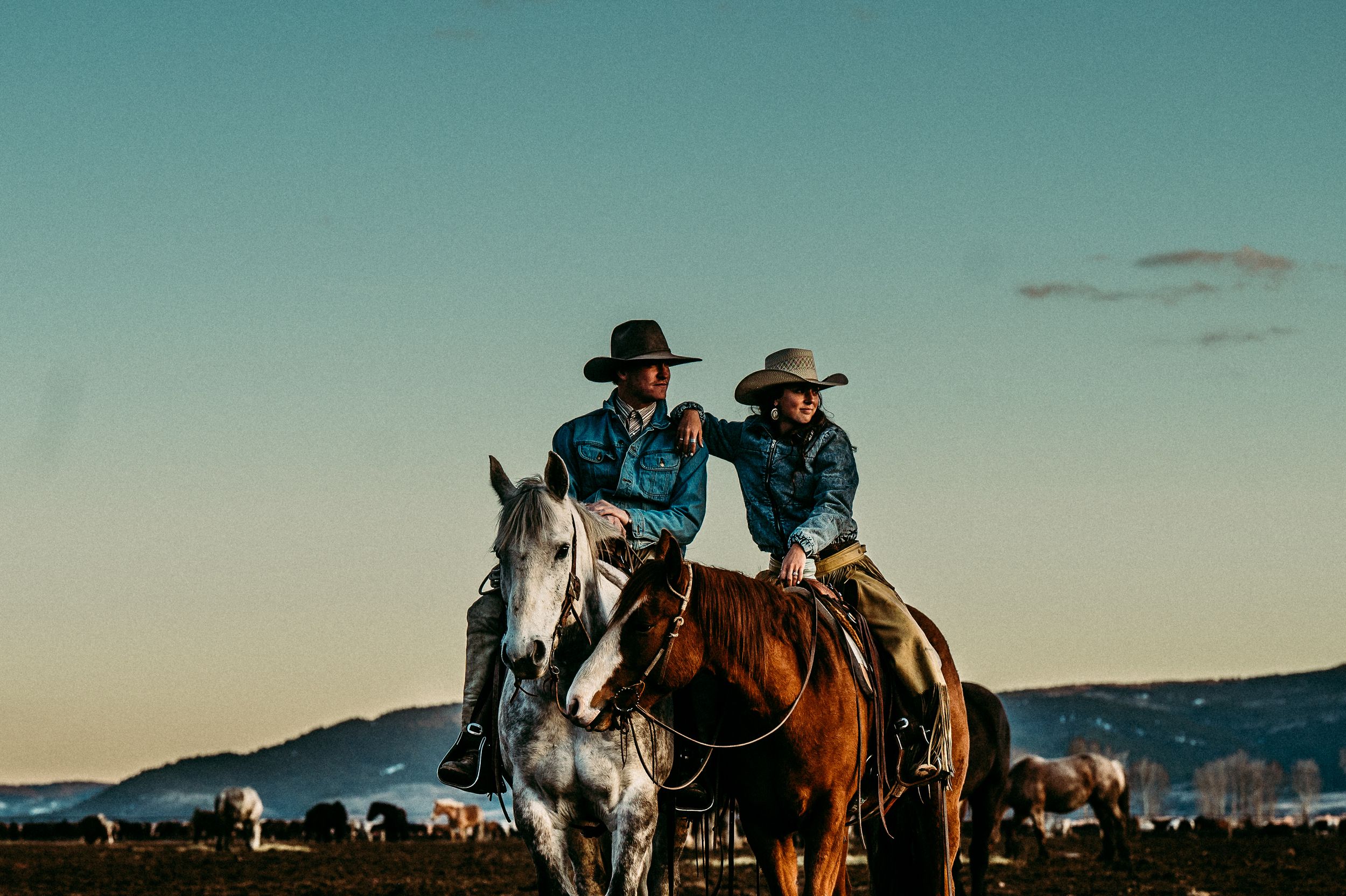 Two cowboys riding horses on plains during sunset with mountains in background against turquoise sky.