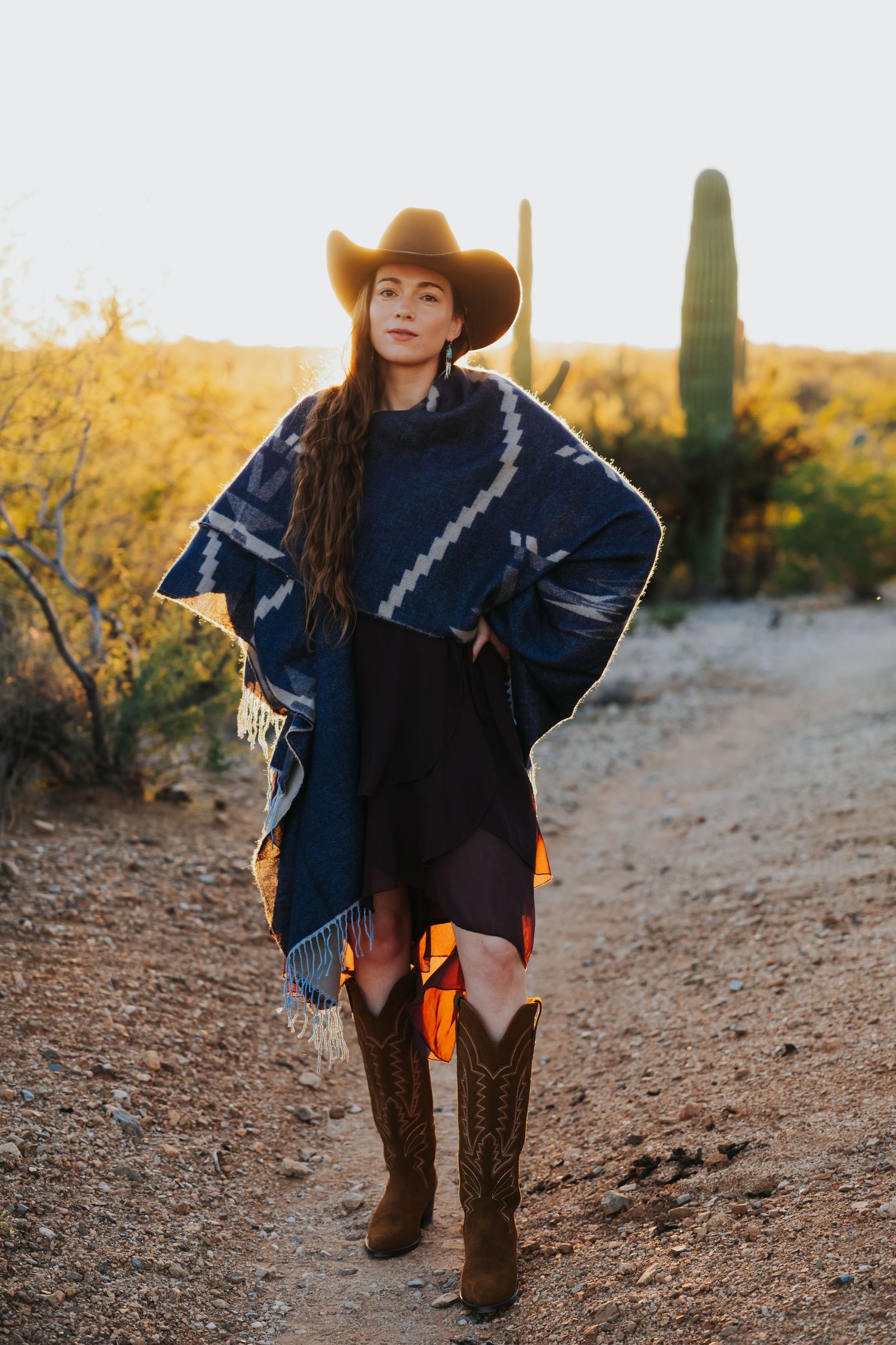 Woman in western attire with cowboy hat and boots poses in desert landscape with saguaro cactus.