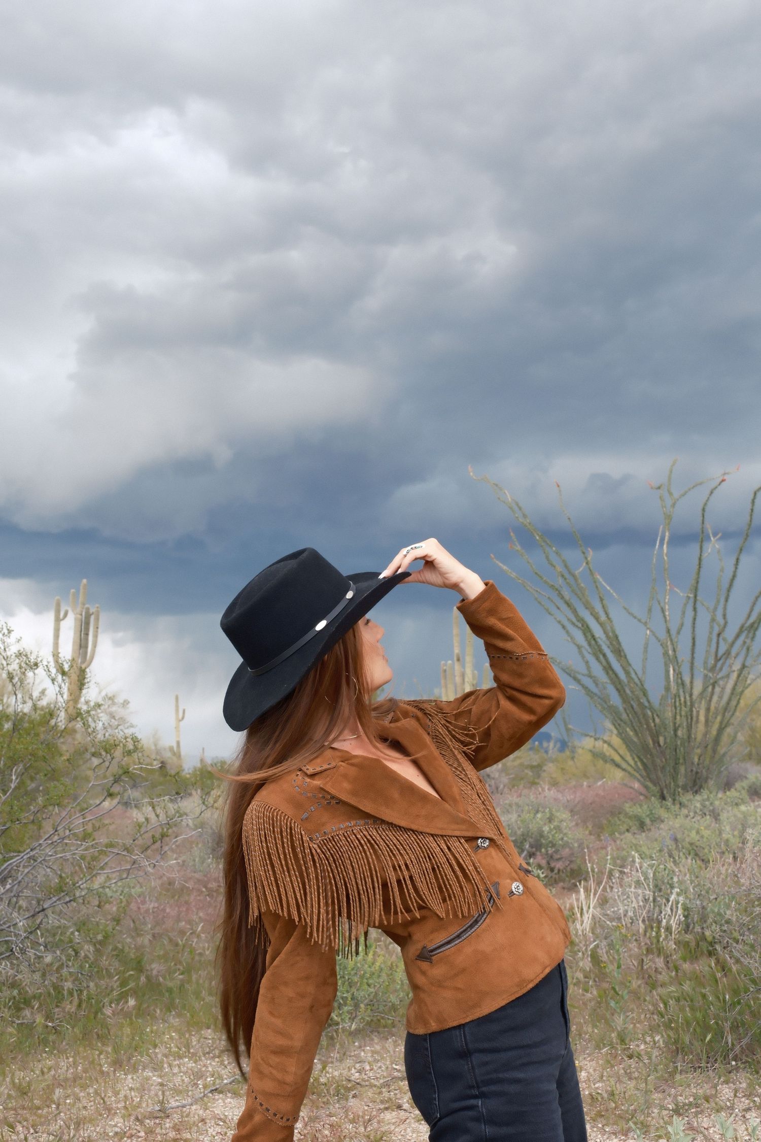 Person in fringe suede jacket and black hat poses against desert landscape with stormy sky.