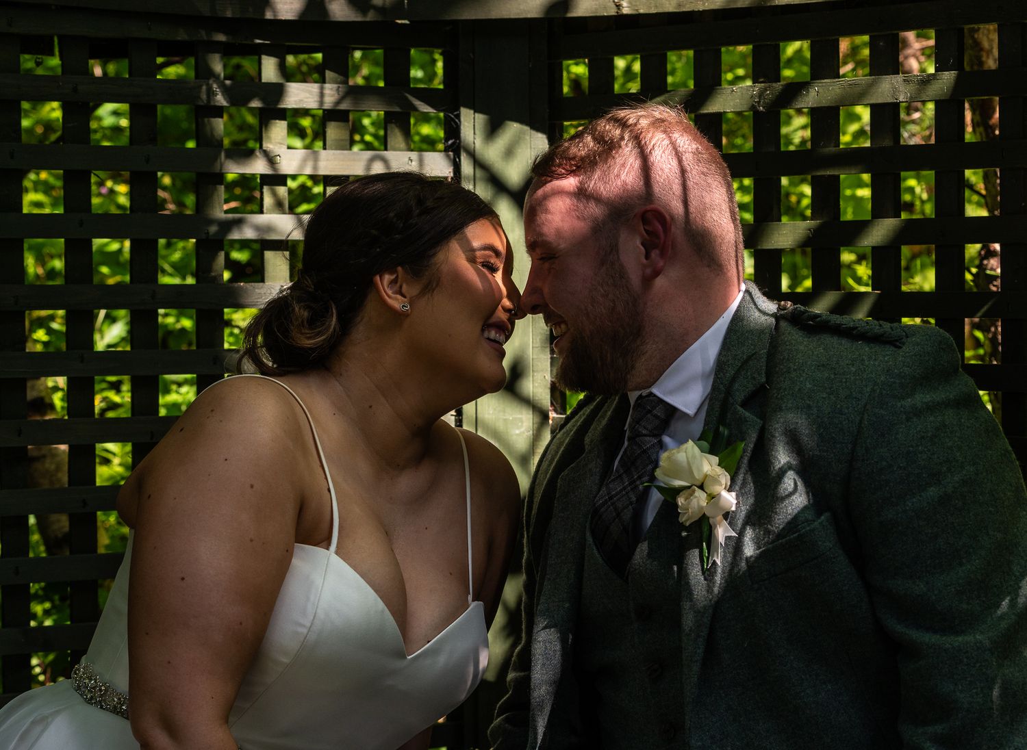 A romantic wedding couple shares a tender kiss against a green latticed backdrop decorated with foliage.