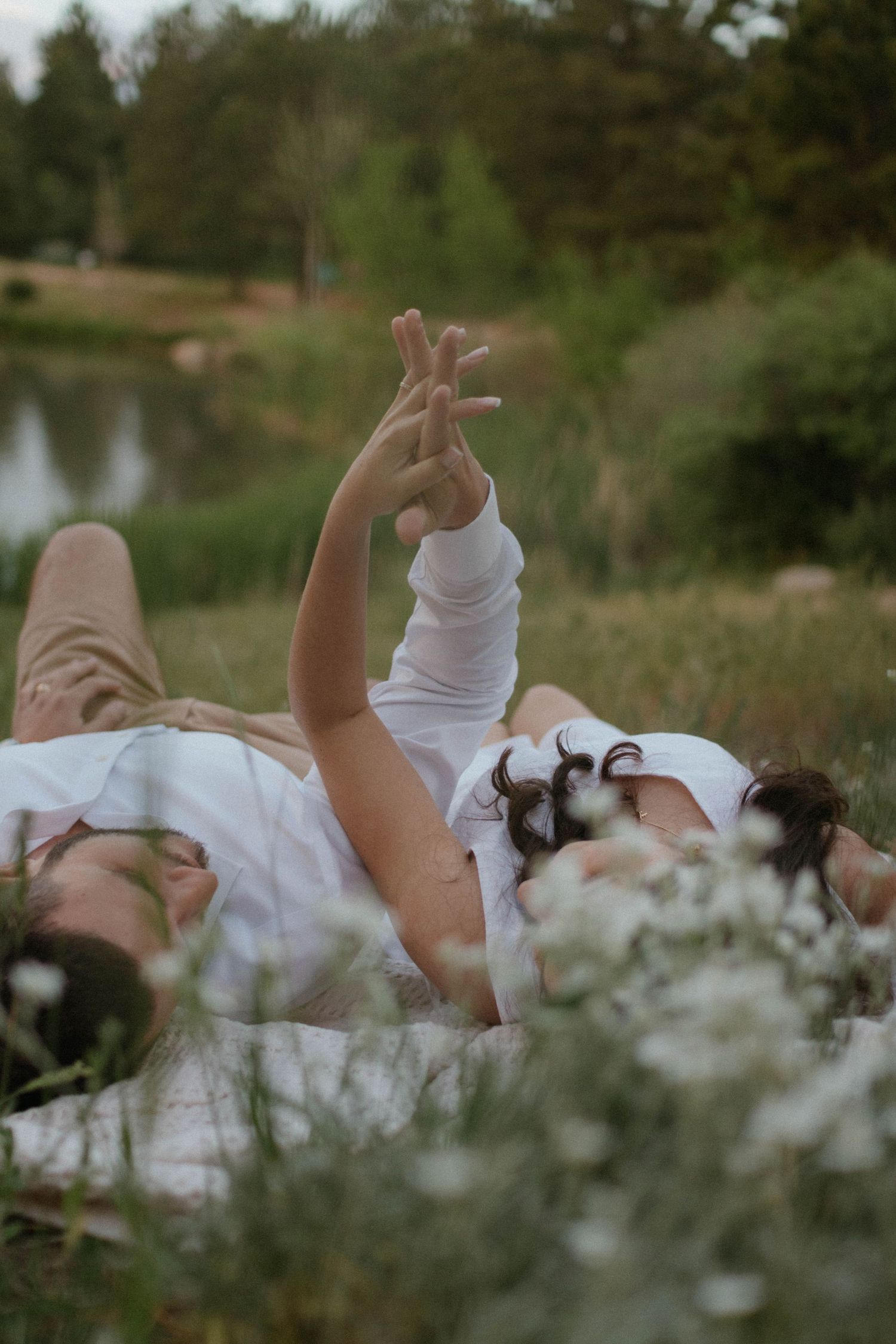 A dreamy summer scene with a couple lying in the grass by a pond, with their arm raised gracefully against a blurred background. Engagement