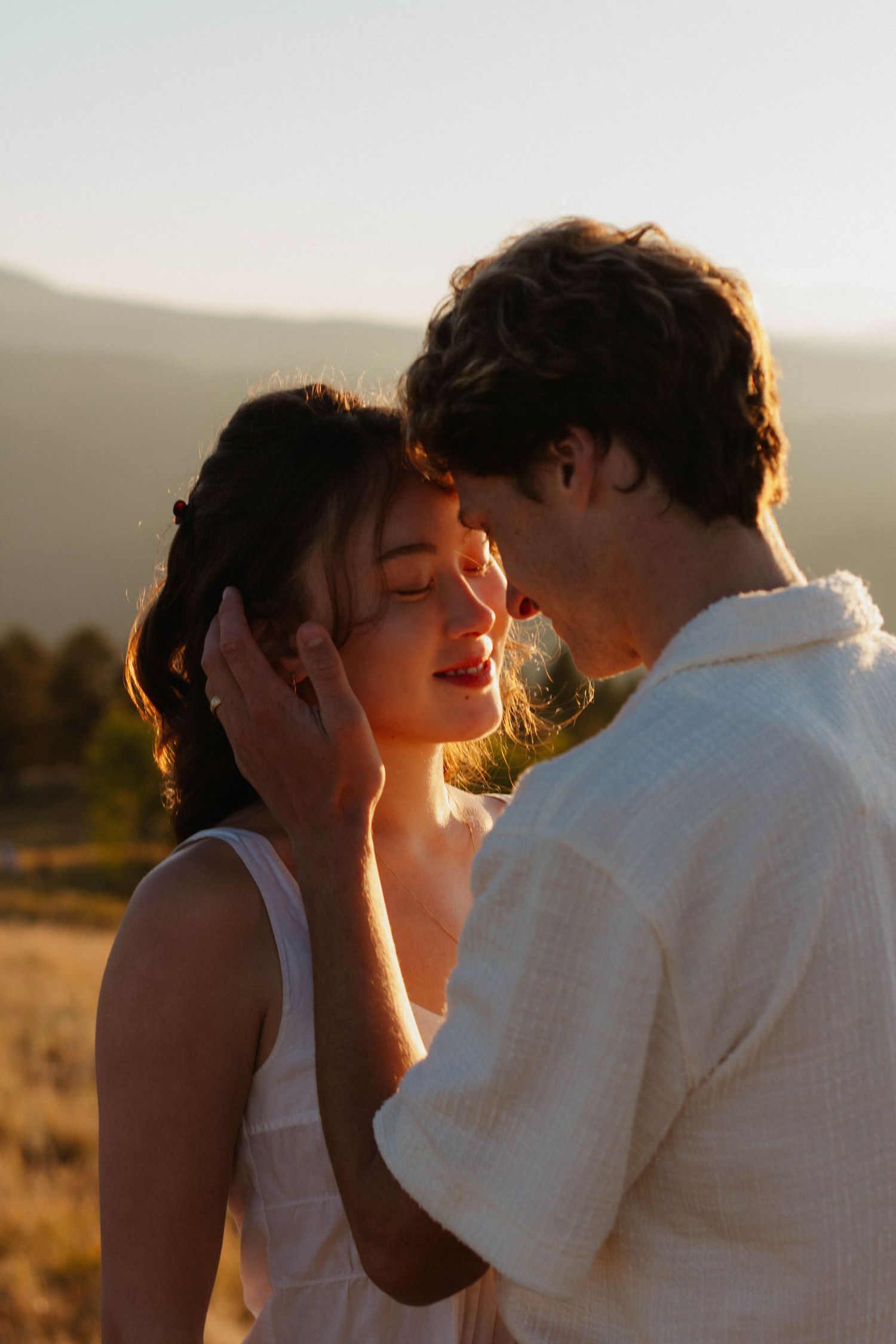 A romantic couple embraces and kisses during golden hour with sunlight creating a warm backlit silhouette.