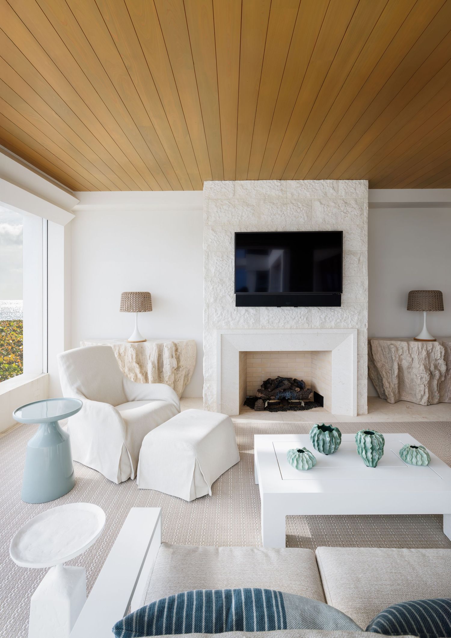 Bright coastal living room with wood ceiling, white fireplace, TV, and teal accents.