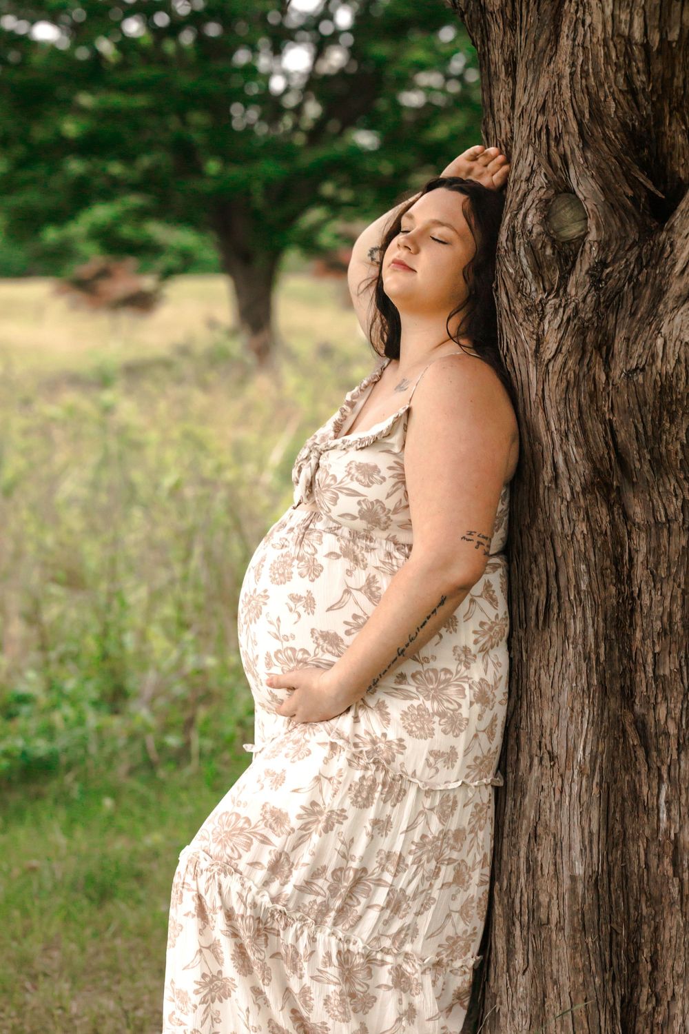 Woman in a floral dress poses against a rustic tree trunk in a natural outdoor setting.