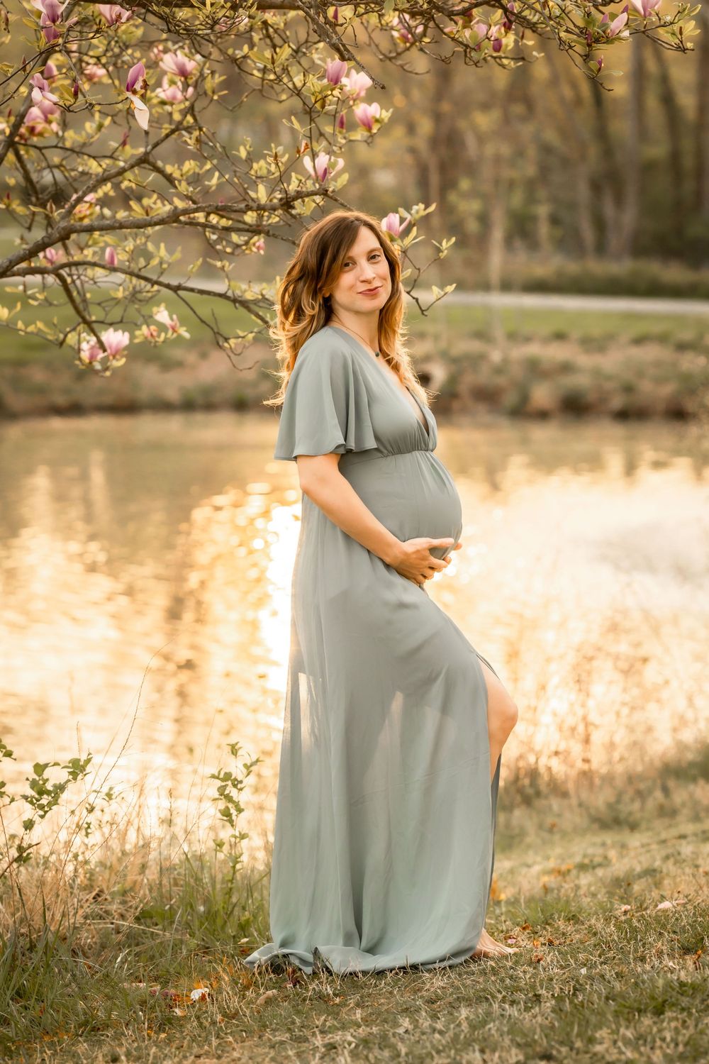 An expectant mother in a flowing sage dress poses by a pond under blooming spring trees. Annapolis maternity photographer
