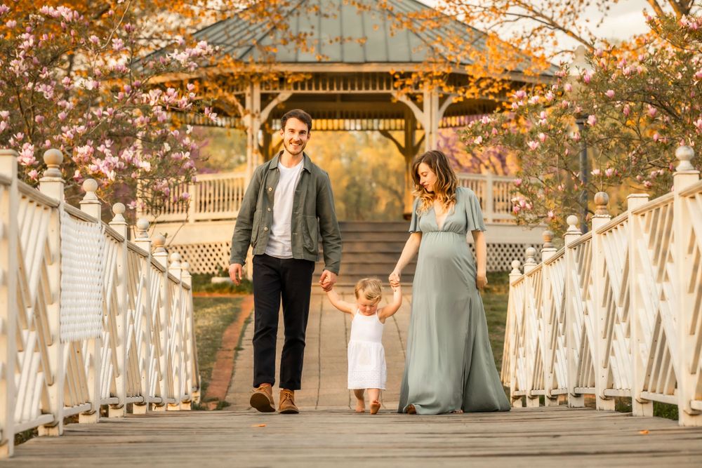 A family walks together on a wooden bridge with a gazebo in the background during golden hour. Annapolis maternity photography studio