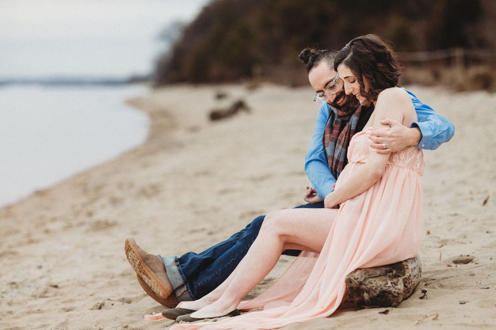 A couple sits together on a sandy beach during a romantic photoshoot. Annapolis maternity photographer. Edgewater photographer