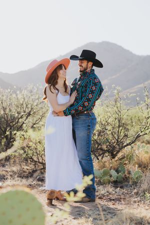 A couple embraces while standing in a desert landscape with mountains in the distance.