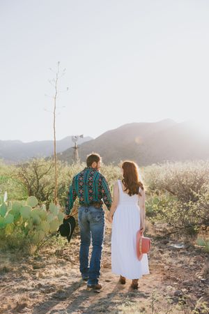 A couple walks hand in hand through a desert landscape at sunset wearing western-inspired outfits.