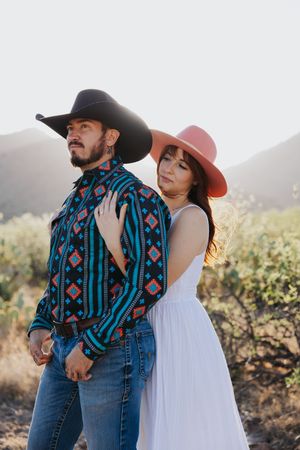 A couple poses together in western attire with mountains and desert brush in background.