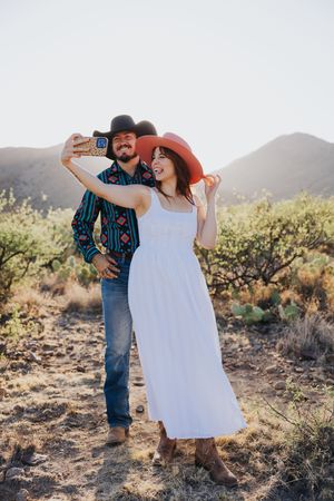 A couple takes a selfie while wearing western hats in a desert setting.