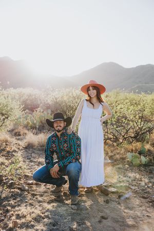 A western-styled couple poses in a desert landscape with mountains and cacti.