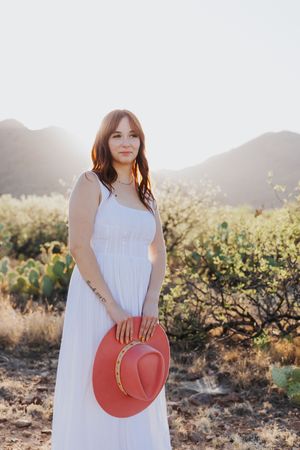 Woman in white dress holds coral hat while posing in desert landscape.