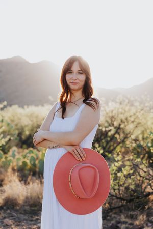 Woman in white sundress poses with coral hat against desert mountain backdrop.