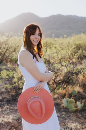 Woman in white dress smiles while holding coral hat in desert setting.