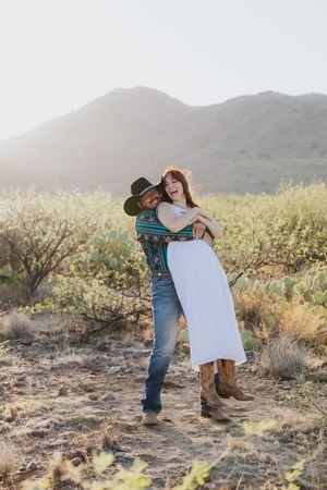 Couple embraces in western wear against desert mountain landscape at sunset.