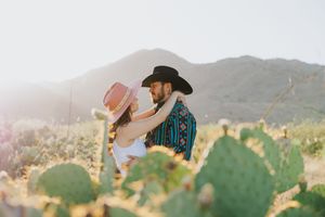 A couple poses together in a desert landscape with mountains and cactus at sunset.