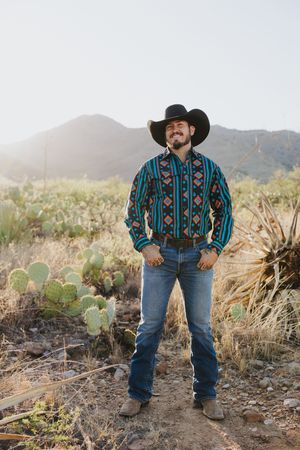 Person in western attire and cowboy hat stands among desert cactus and rocky terrain.