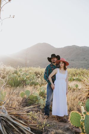 Couple embracing in white dress and western wear among desert cactus at sunset.