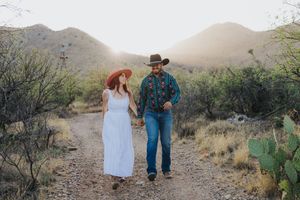 A couple walks hand in hand on a desert trail surrounded by mountains and cacti in the Southwest.