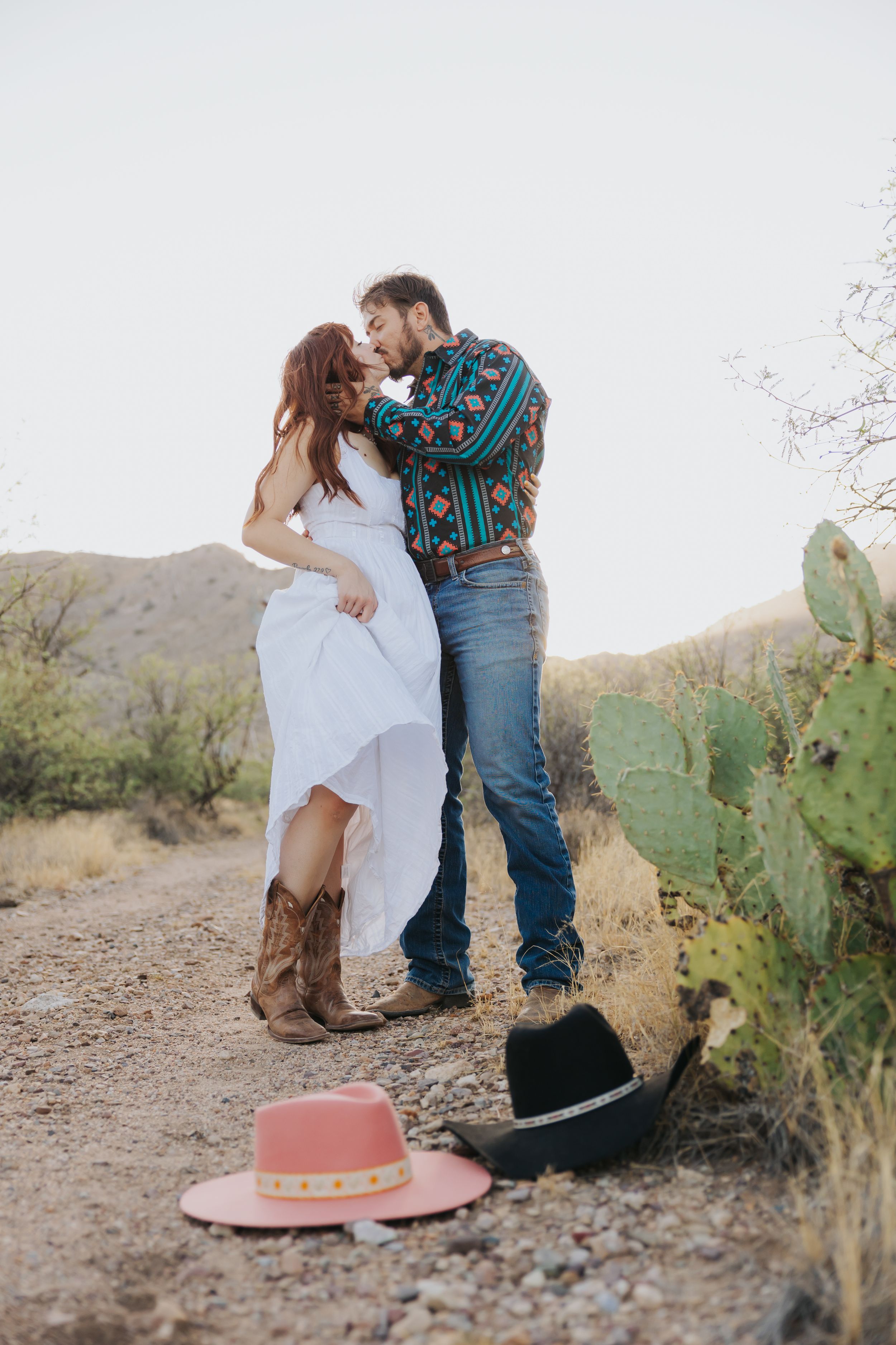 A romantic desert photoshoot with cowboy hats placed on the ground and cacti in the background.