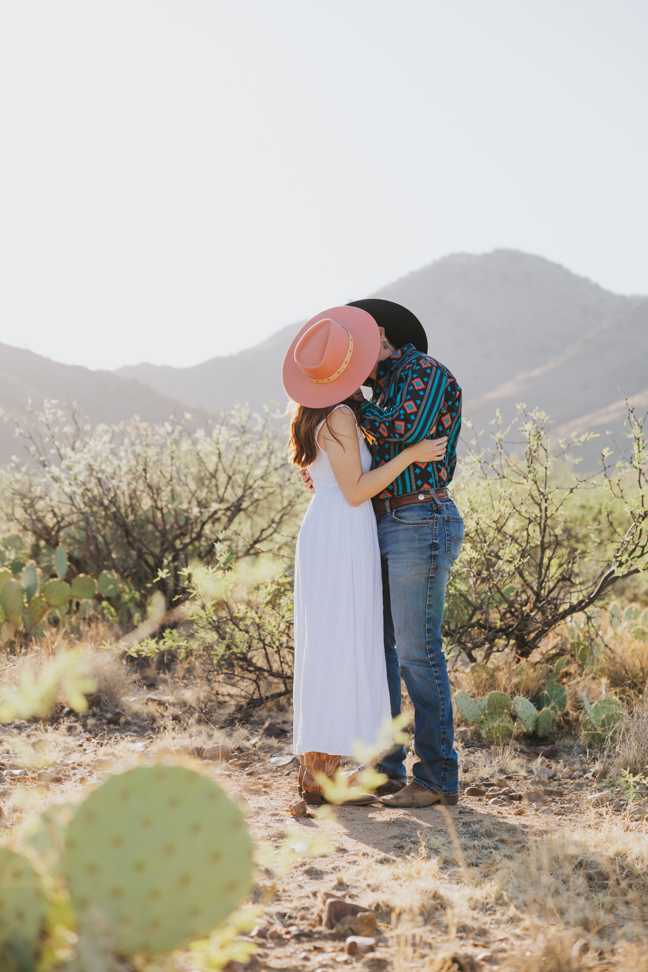 Two people share an intimate moment among desert plants and mountain scenery.