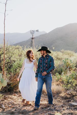 A couple in western attire walks together through a desert landscape with cacti.