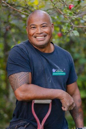Gardener in navy shirt with tribal tattoo holding garden tools among fruit trees.