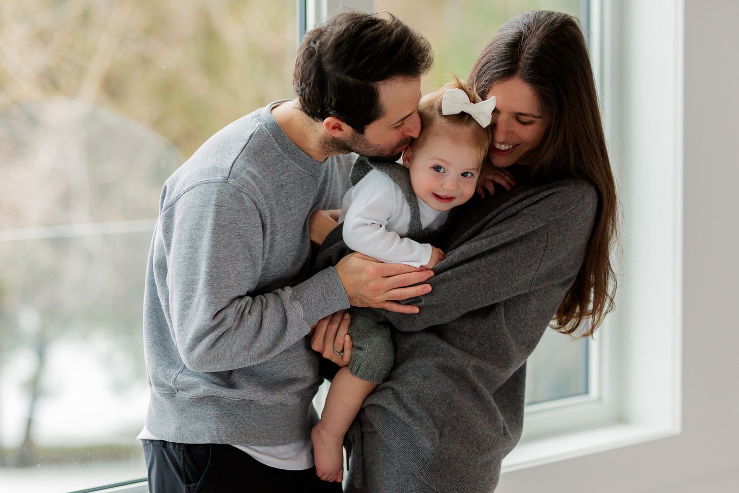 In home family photography session. Family wearing coordinating grey sweaters hugging by a bright window in a cozy indoor setting.