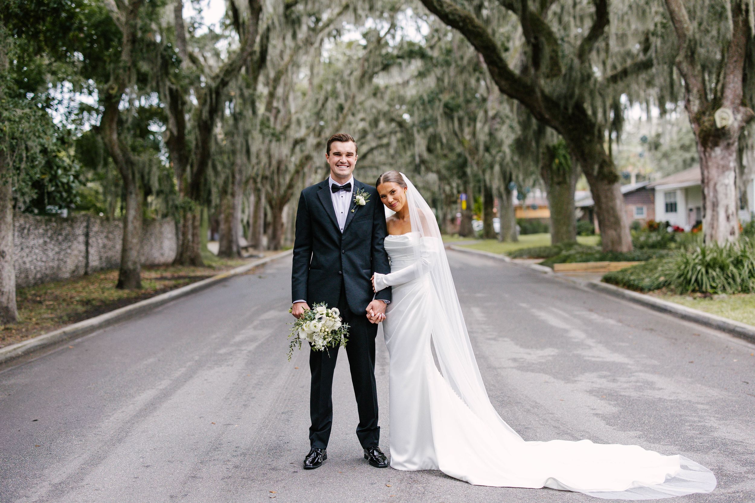 A couple in wedding attire poses under a canopy of Spanish moss draped oak trees along a scenic road.