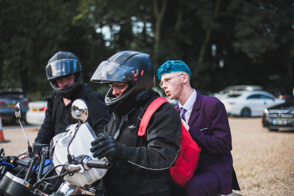 Three people by motorcycles, with one wearing a distinctive purple suit and blue cap.