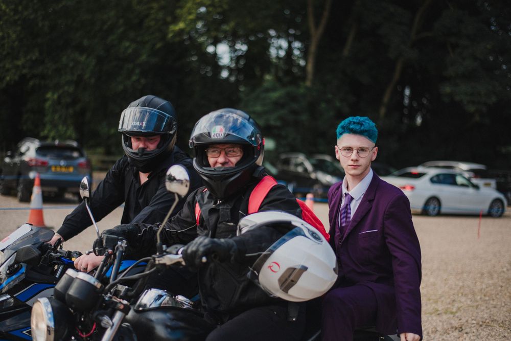 Motorcyclists in helmets and protective gear joined by someone in a purple suit outdoors.