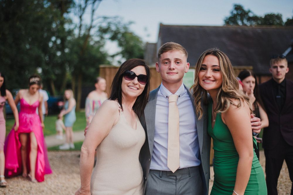 Three guests in formal attire smile together at an outdoor celebration with a barn in the background.