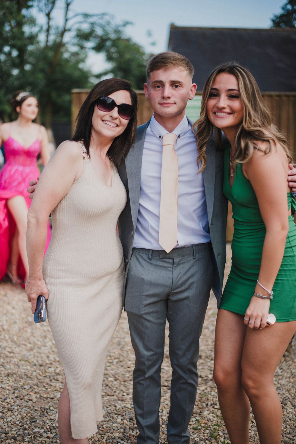 Three guests pose together at an outdoor event, wearing formal attire in neutral and green tones.