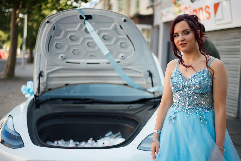 A prom attendee in a sparkling blue ball gown poses next to a white car with an open hood.