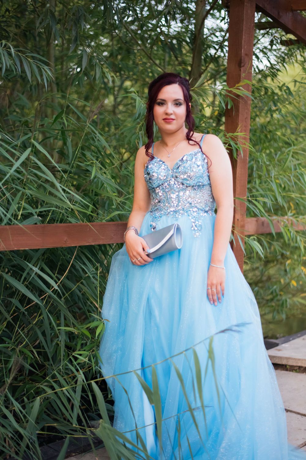Someone in a light blue beaded gown stands on a wooden deck surrounded by greenery.