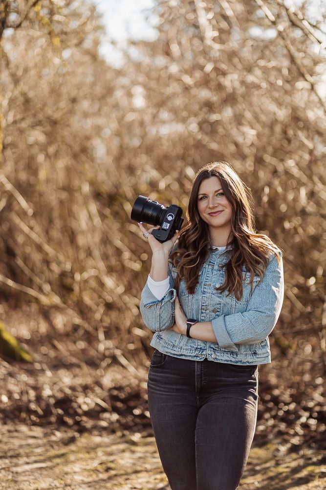 A person in a denim jacket and dark jeans poses outdoors in autumn sunlight against a natural wooded backdrop.