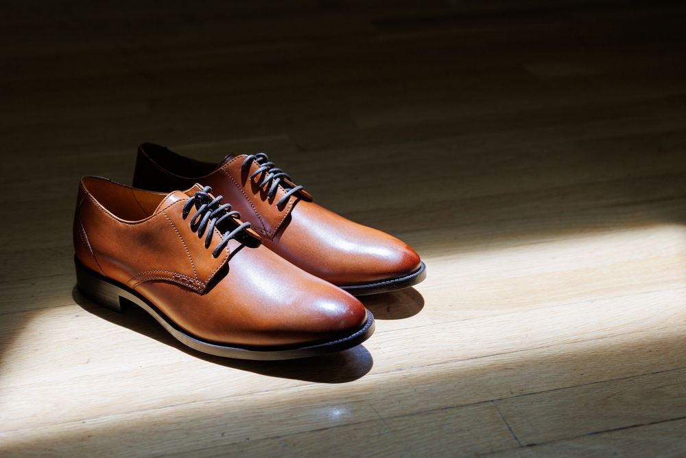 A pair of tan brown leather Oxford dress shoes with dark laces on a wooden floor in dramatic lighting.