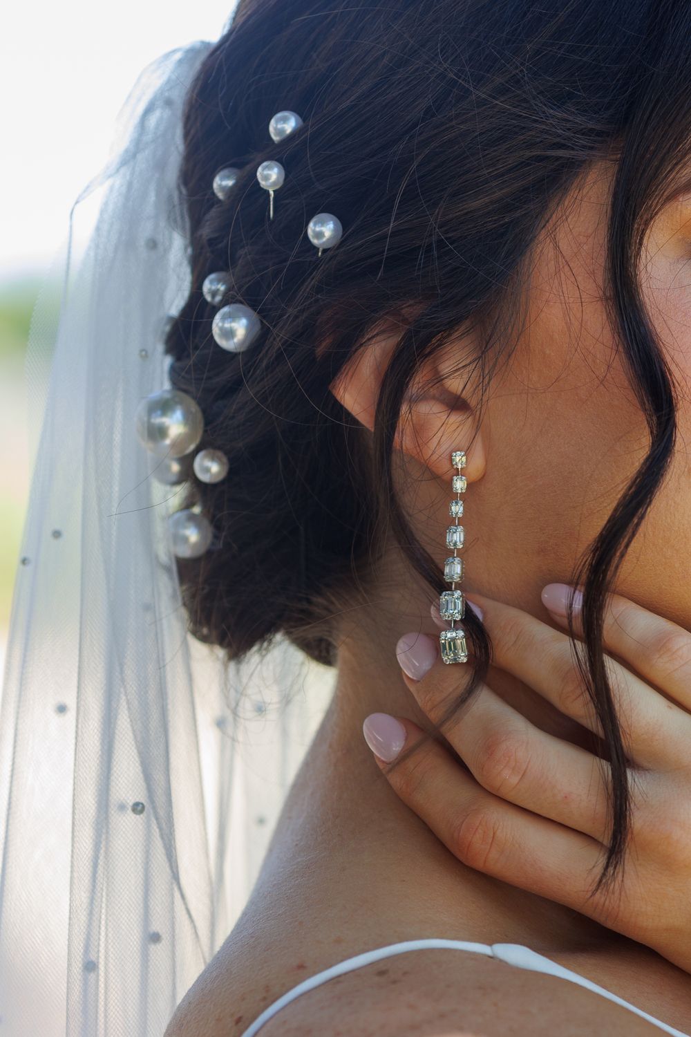 Bride with dark updo hair adorned with pearl pins, wearing elegant crystal drop earrings and a white veil.