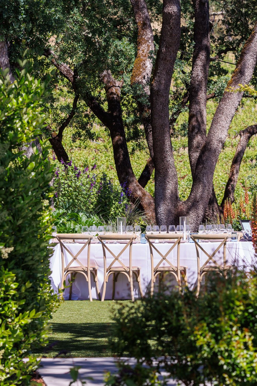 Elegant outdoor garden dining table set with white linens and wooden cross-back chairs beneath a large oak tree.