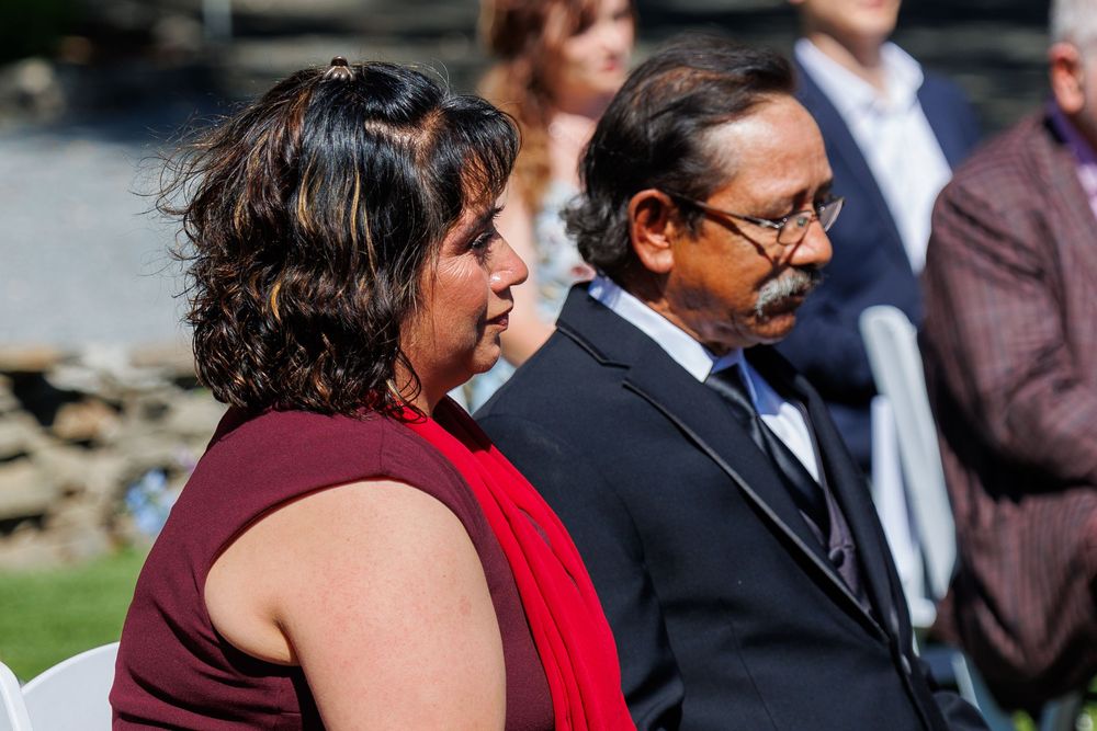 Two guests in formal attire seated outdoors at a wedding ceremony on a sunny day.