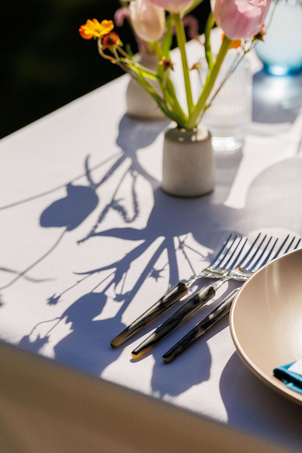 Elegant outdoor table setting with cutlery, ceramic plate, and fresh flowers casting artistic shadows on white tablecloth.
