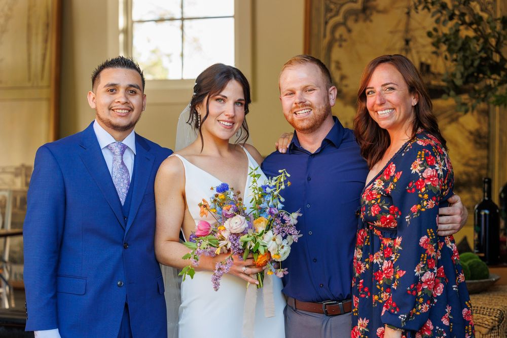 Wedding couple with guests posing indoors, bride holding colorful bouquet, groom in blue suit, smiling group photo.