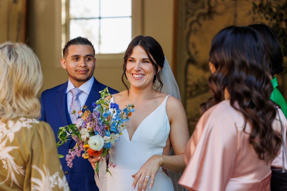 Smiling bride holding colorful bouquet stands with groom in blue suit during wedding ceremony greeting guests.