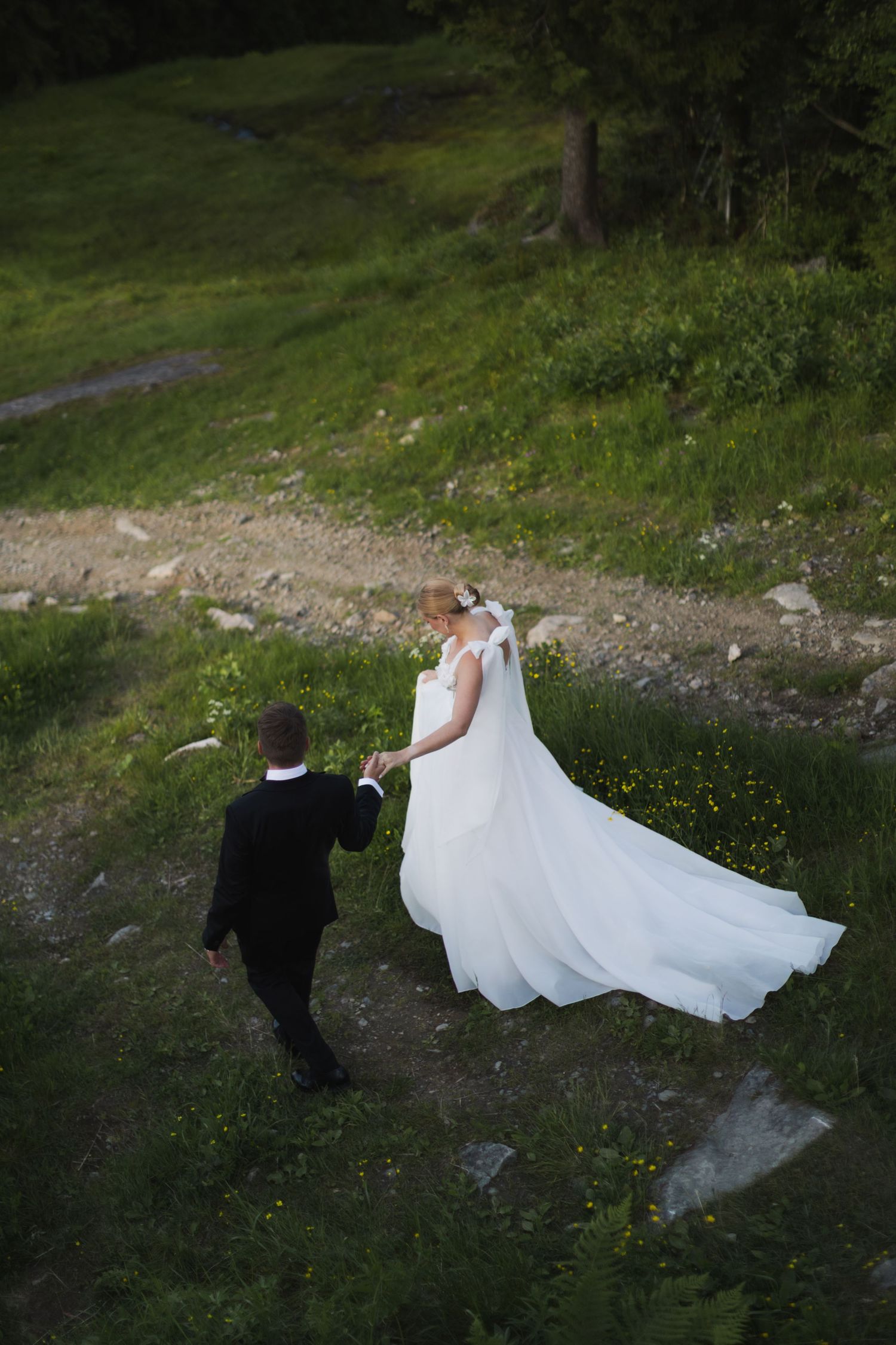Bride in white gown and groom in black suit walk hand in hand through lush green hillside landscape.