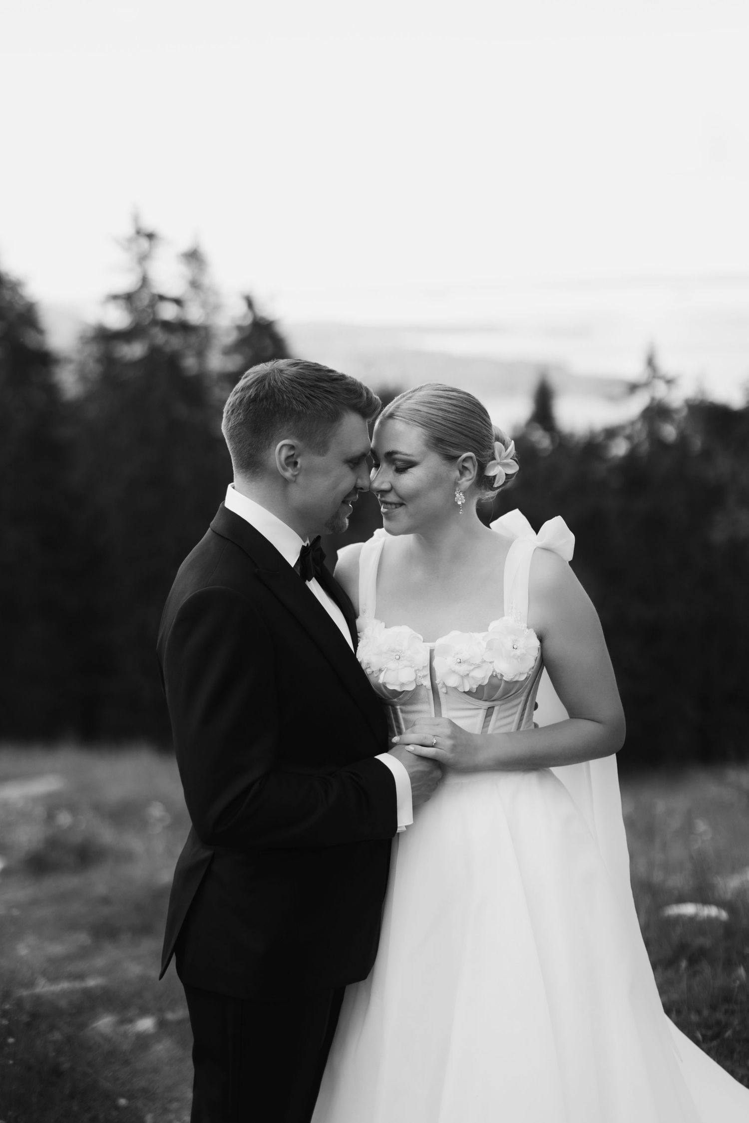Black and white photo of a bride and groom in formal attire sharing an intimate moment outdoors near evergreen trees.