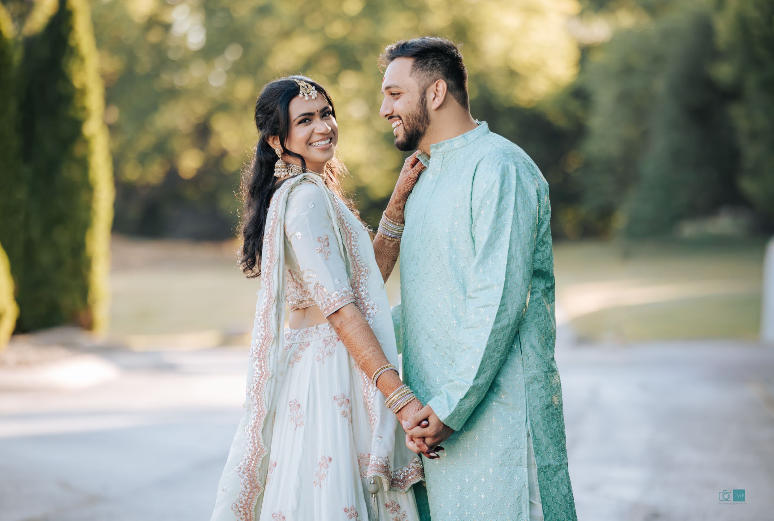 Indian couple in traditional wedding attire, bride in white lehenga and groom in mint kurta, posing outdoors.
