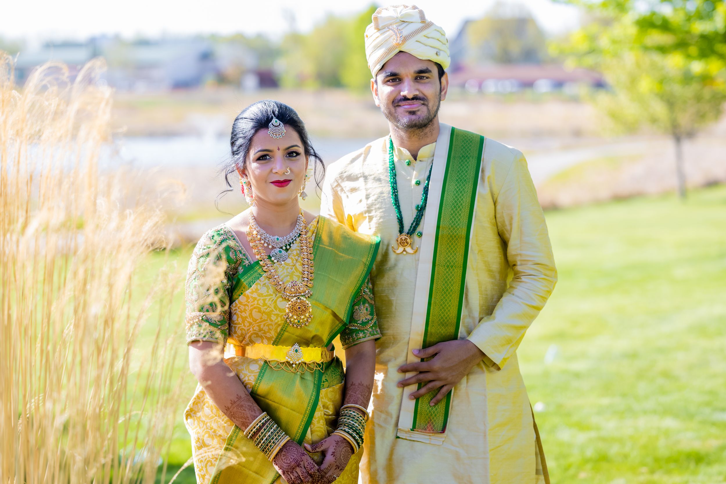 Indian couple in traditional wedding attire, bride in green saree and groom in gold sherwani, posing outdoors.